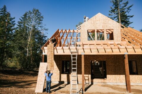 A wooden house under construction with workers installing plywood sheathing on the roof and walls.